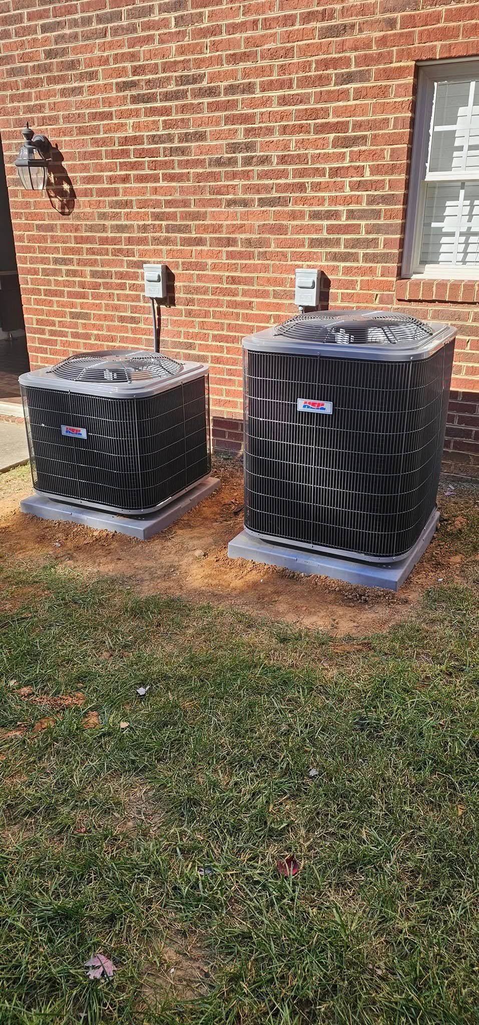Two black air conditioning units on concrete pads in front of a brick wall and a window. They are on a patch of grass.