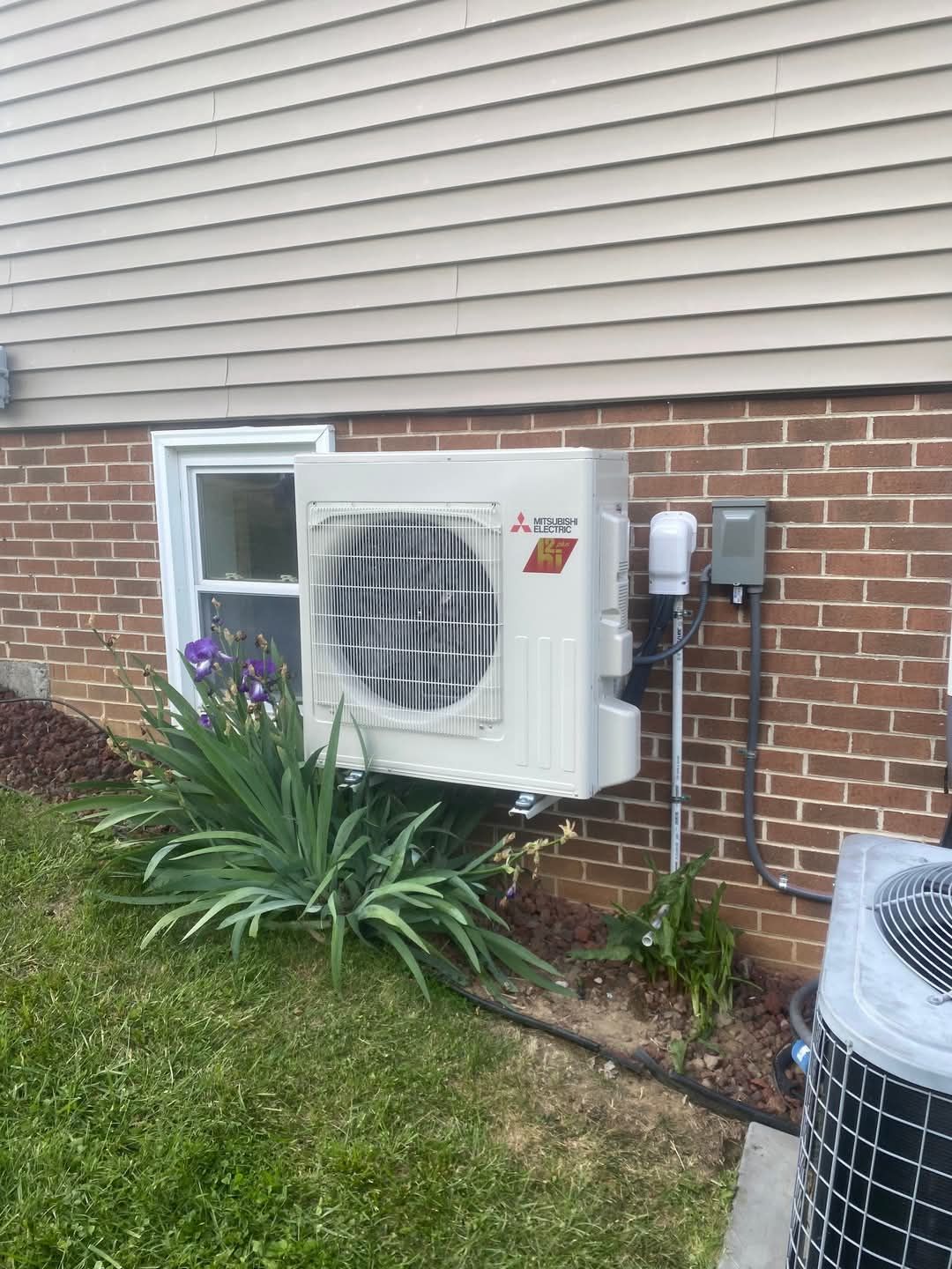 An air conditioning unit is mounted on a brick wall next to a small window. Green plants grow in the foreground.