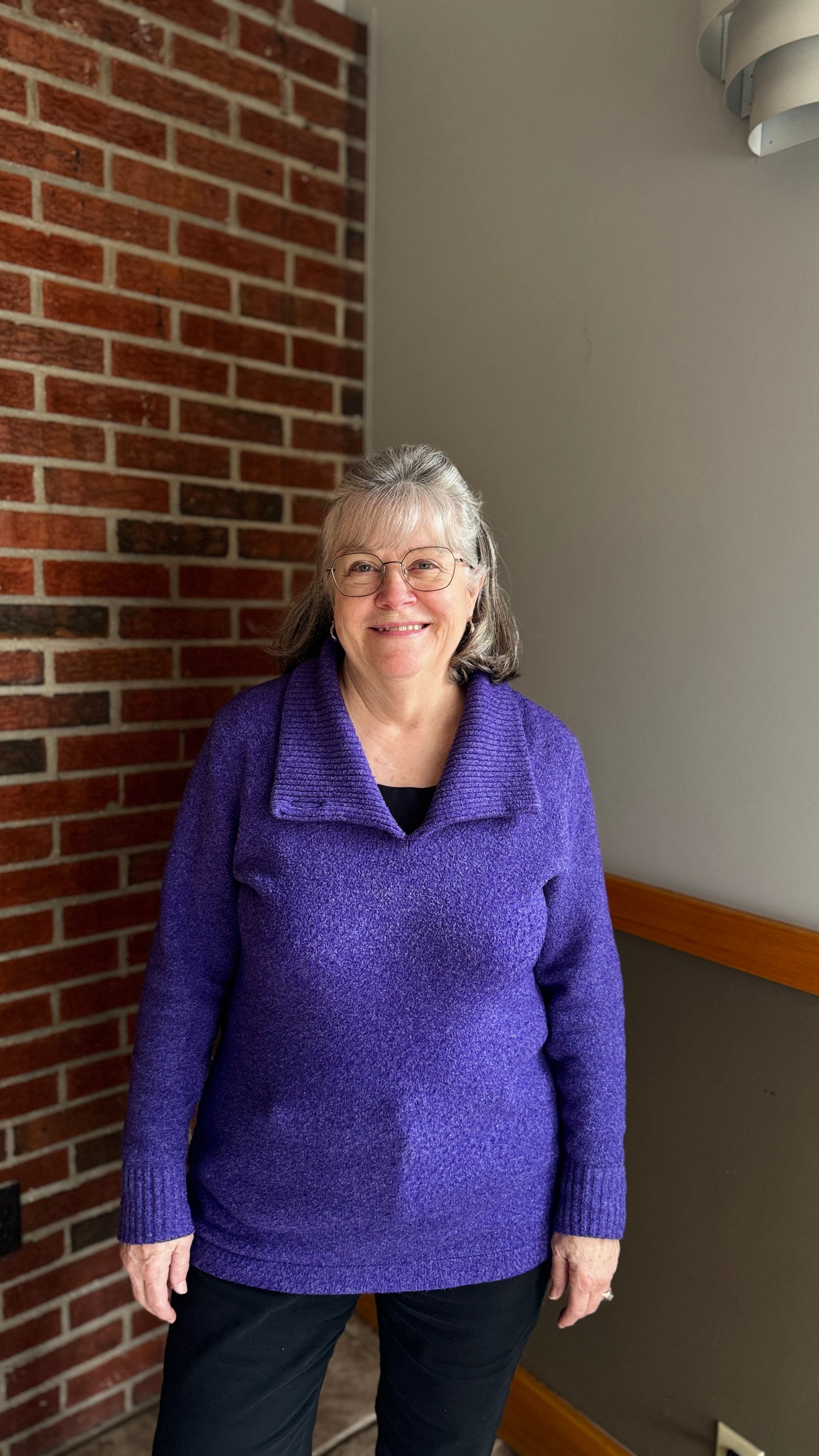 A woman wearing glasses and a blue shirt is smiling in front of a brick wall.