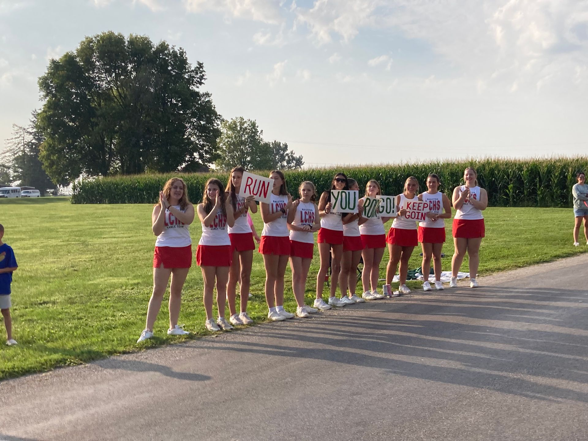 A group of cheerleaders are standing on the side of the road holding signs.