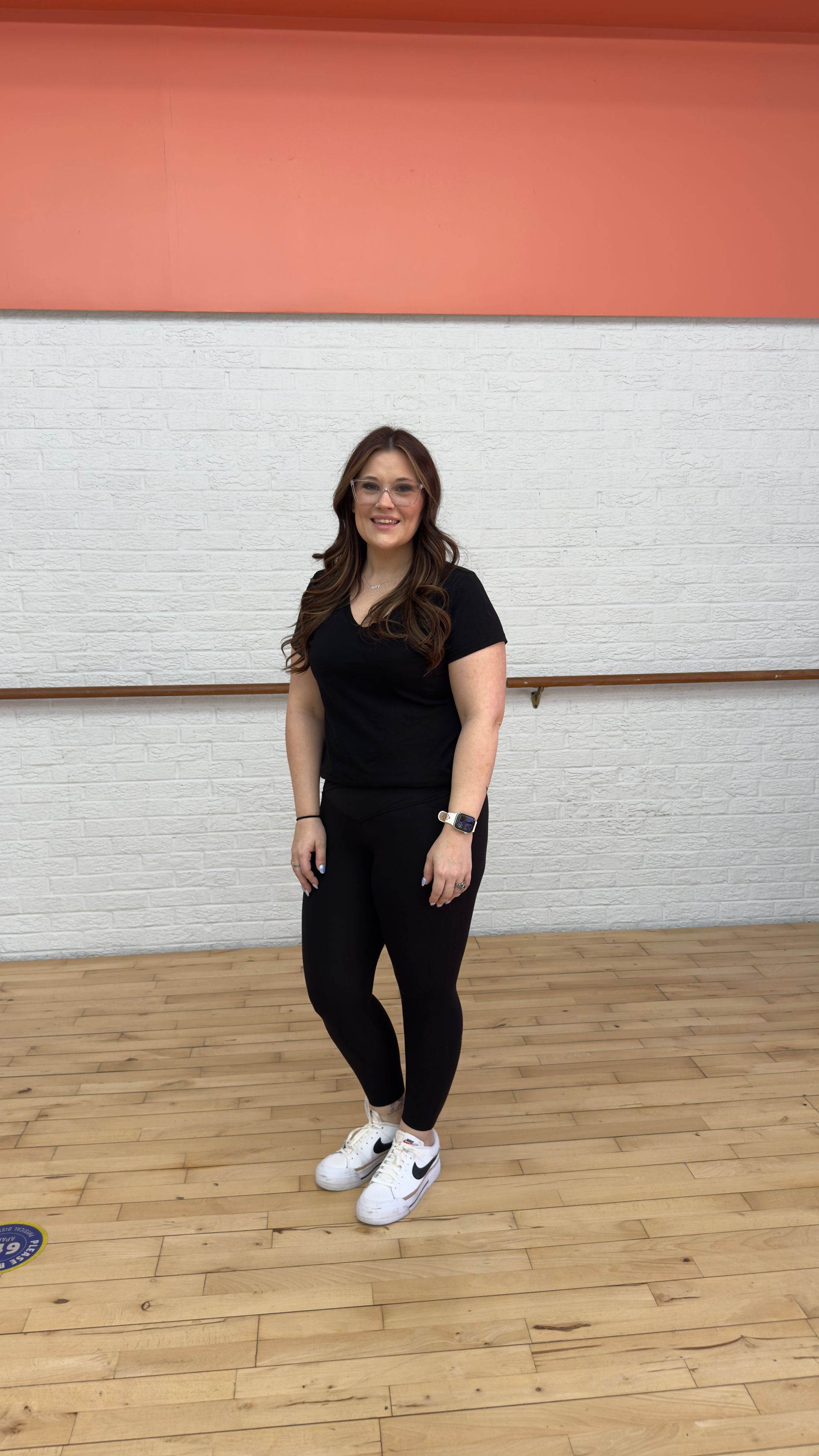 A woman in a black shirt and white sneakers is standing in a dance studio.