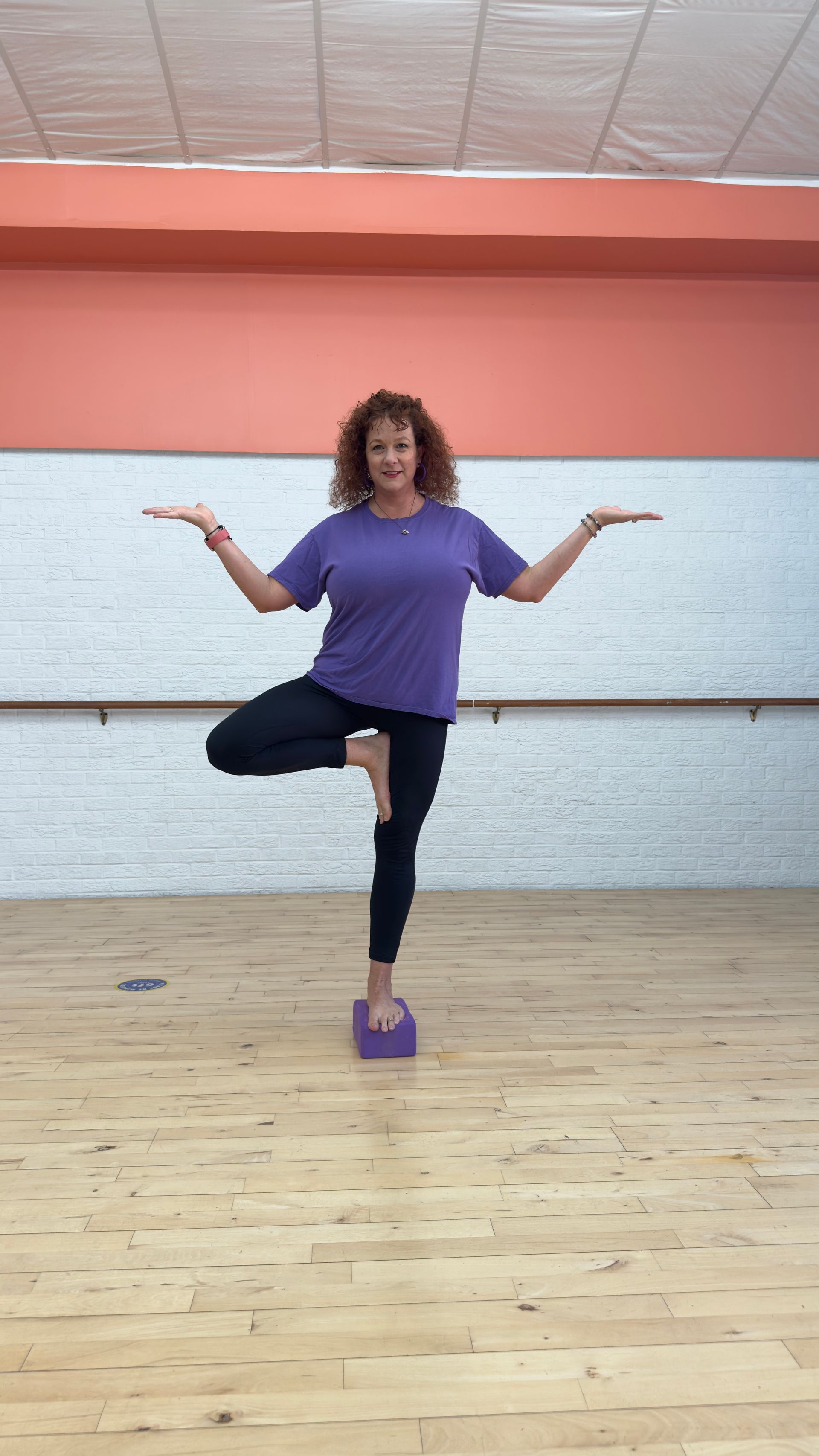 A woman is standing on one leg on a yoga block in a dance studio.