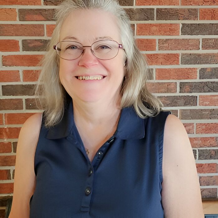 A woman wearing glasses and a blue shirt is smiling in front of a brick wall.