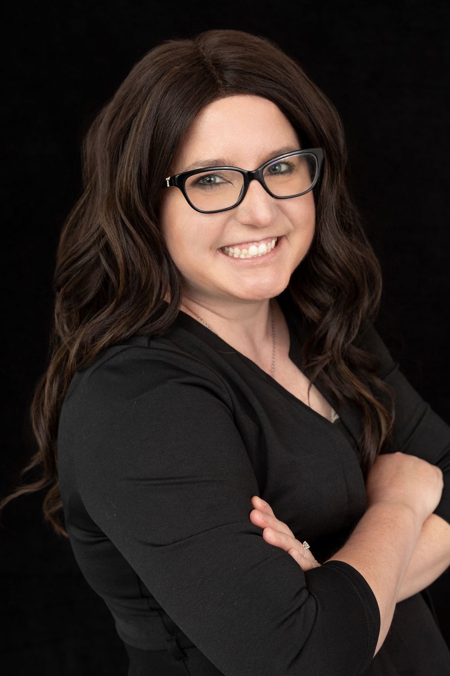 Smiling person with dark hair and glasses, arms crossed, wearing a black top against a dark background