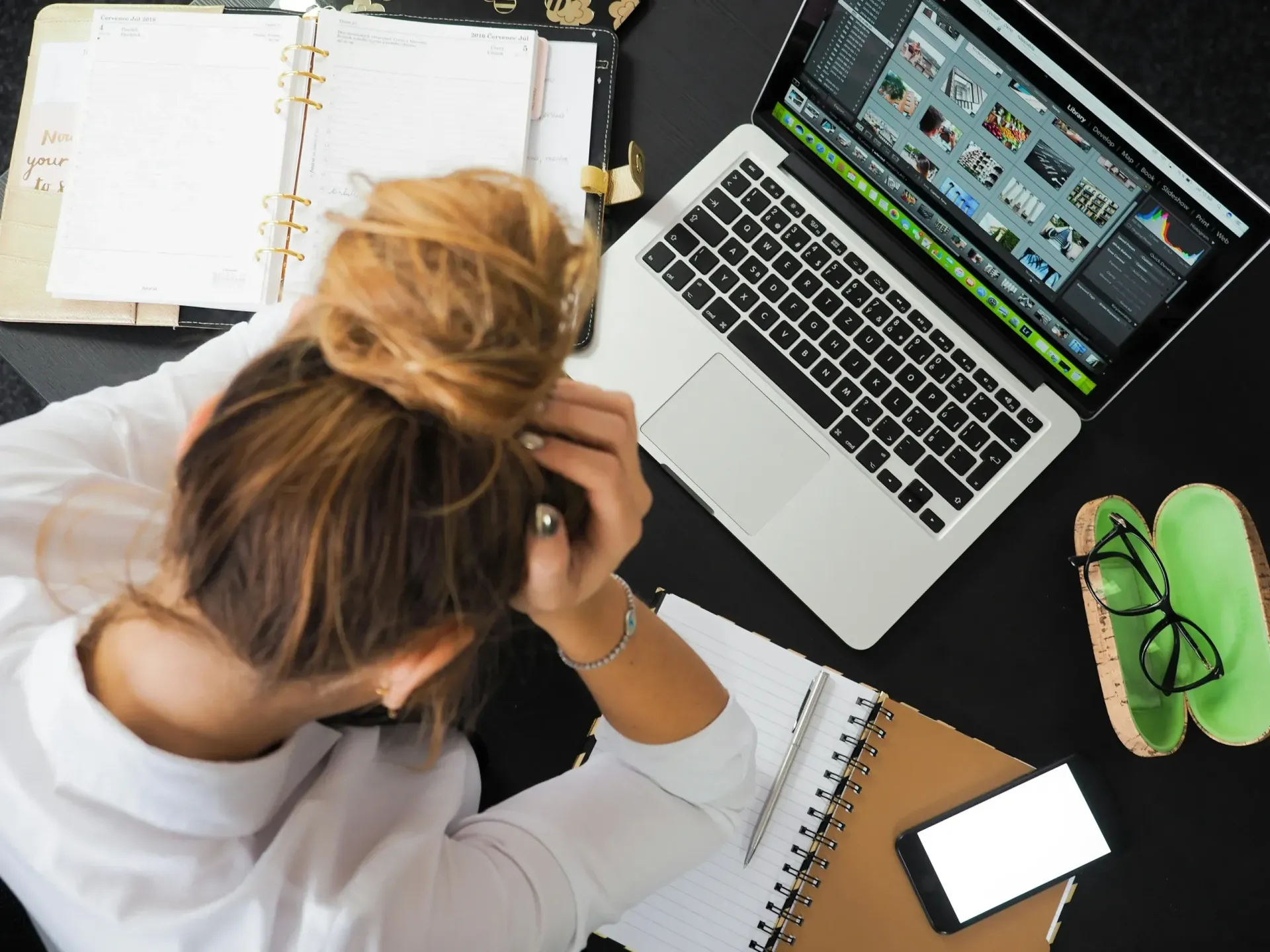 Person working at a desk with a laptop, notebooks, phone, and green glasses.