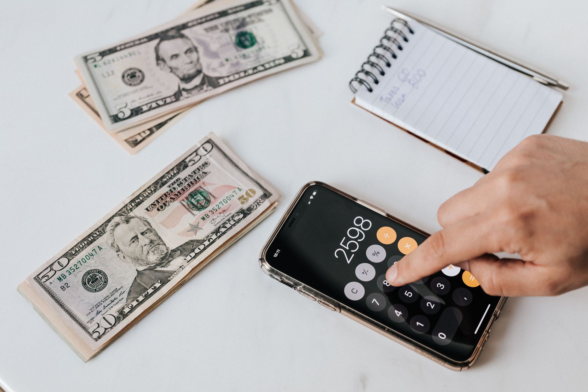Hand entering numbers on calculator beside cash and notebook on a white desk