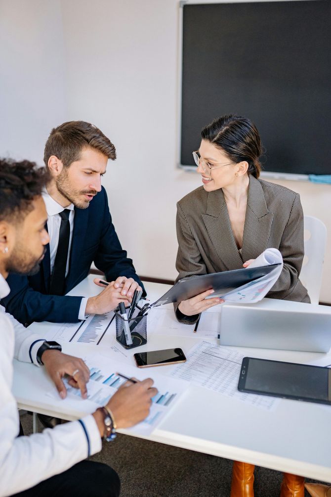 Three coworkers in a meeting, reviewing documents around a laptop at a white conference table.