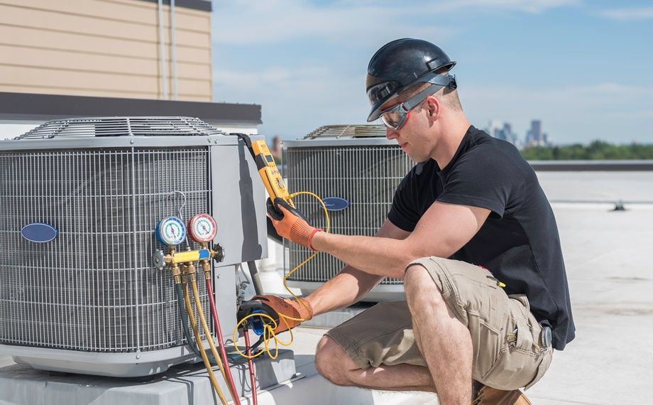 HVAC technician kneeling near AC unit, using a meter. Rooftop setting, sunny day.