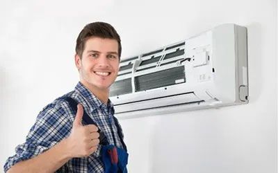 HVAC technician with thumbs up, standing near a wall-mounted air conditioning unit.