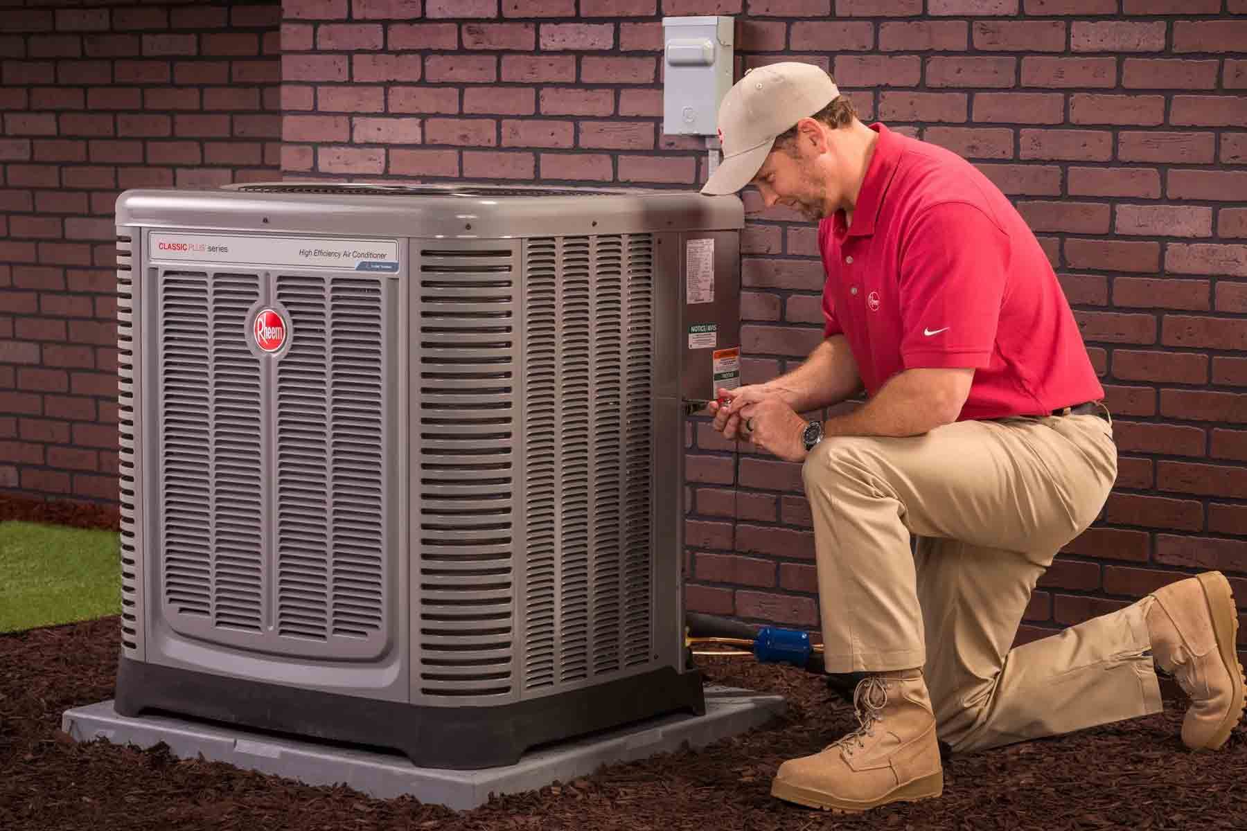 HVAC technician kneels beside air conditioning unit, inspecting it outside a brick building.