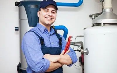 Smiling plumber with crossed arms, holding wrench, standing near water heater and pipes.