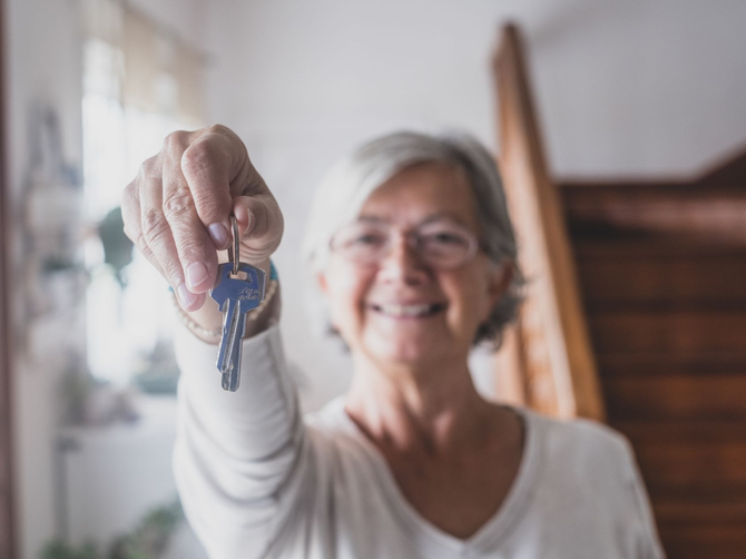 Smiling senior woman holding up house keys near a staircase.