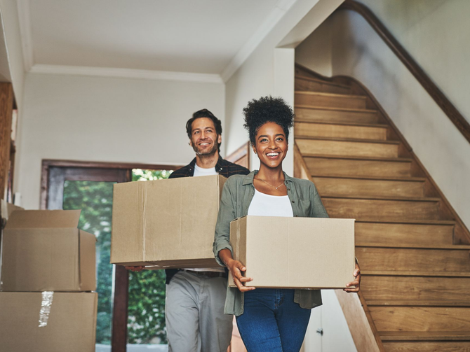 Couple carrying cardboard boxes, smiling, inside a house near a staircase.