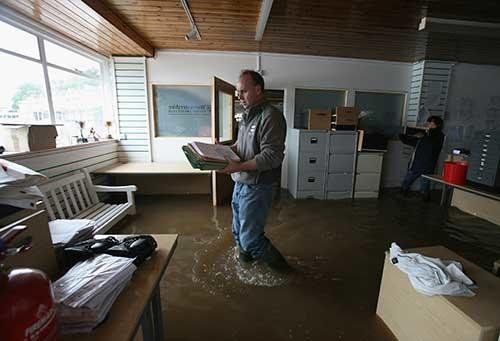 Man wades through flooded office, holding paperwork, as another person films.