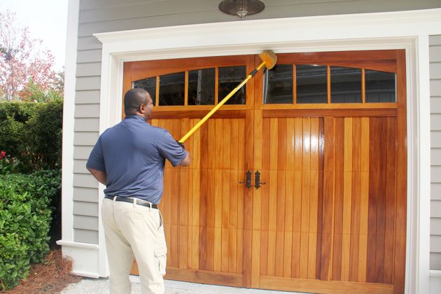 A person using a yellow pole tool to clean or seal the top edge of a wooden garage door.