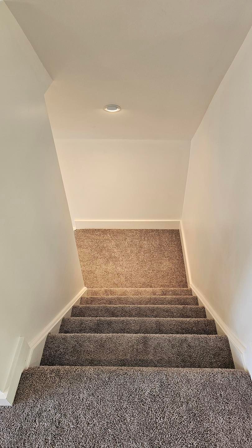 Staircase with gray carpet leading down, white walls and ceiling, single overhead light.