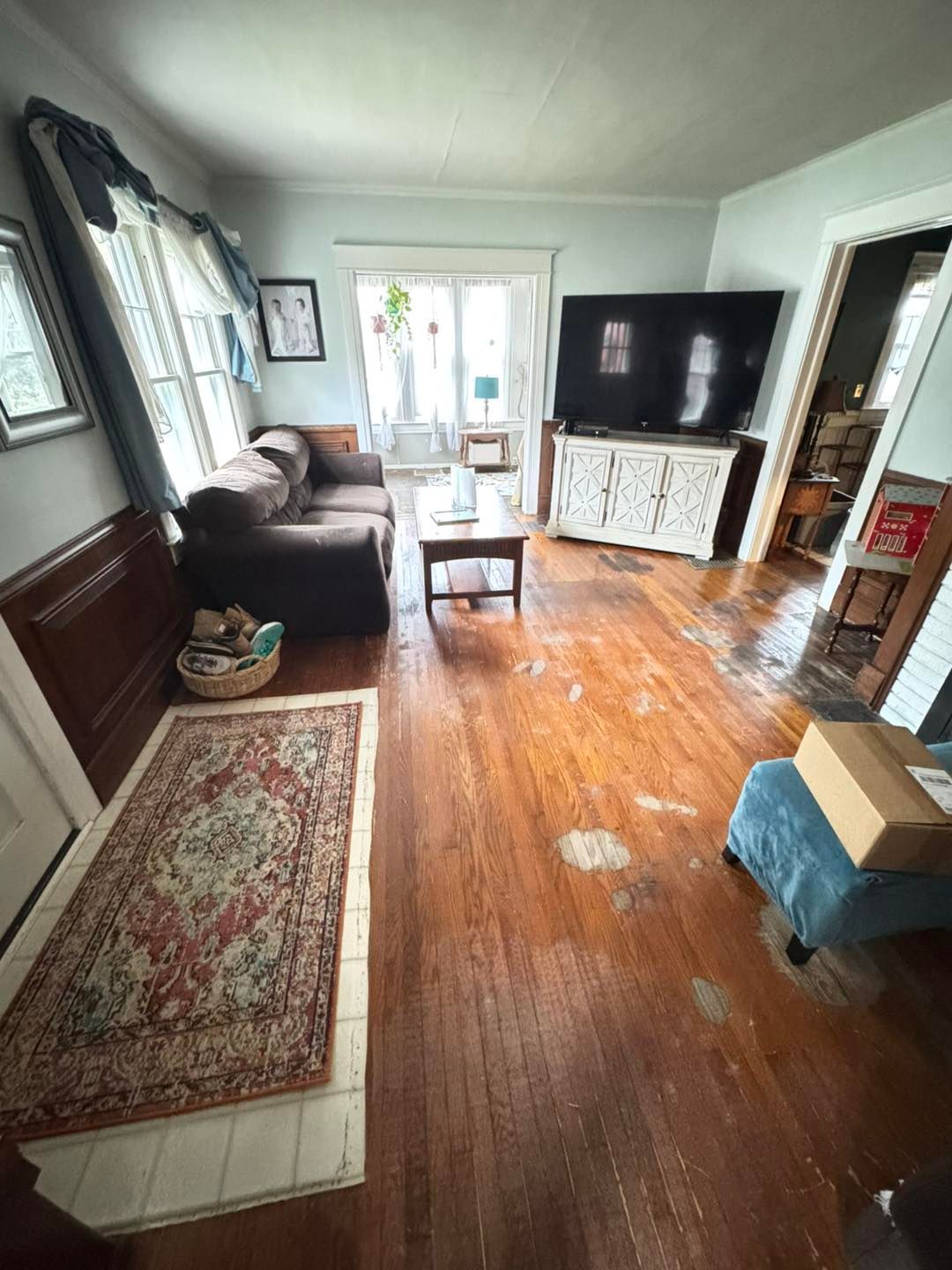 Living room with brown sofa, rug, TV, and wooden floor. Sunlight streams in from a window.