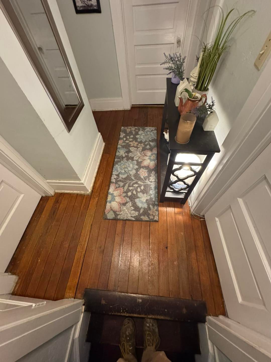 Narrow hallway with wood floors, floral rug, side table with decor, and open door.