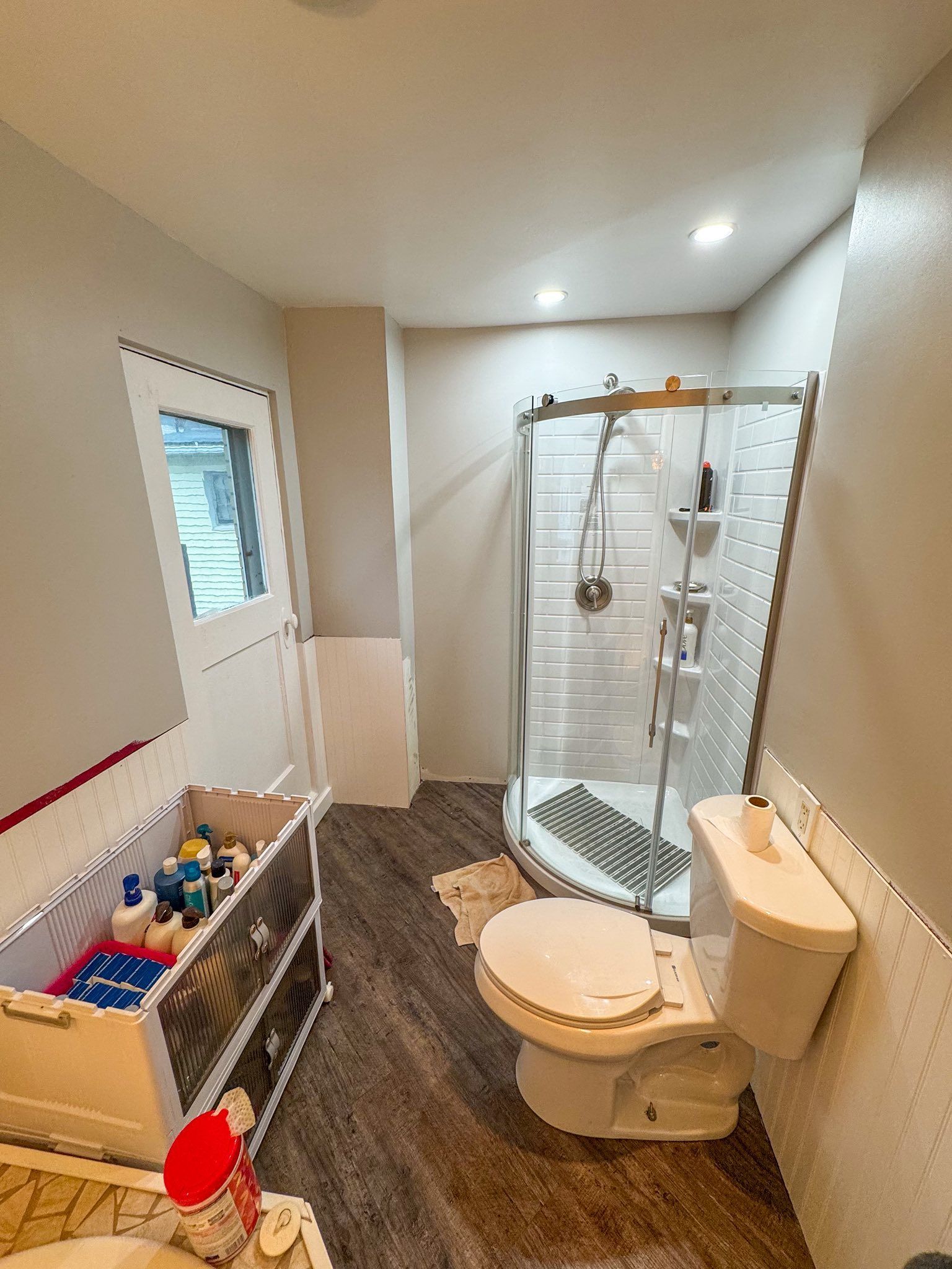 Bathroom with a shower, toilet, and vanity. The walls are light colored, and the floor is a dark wood look.