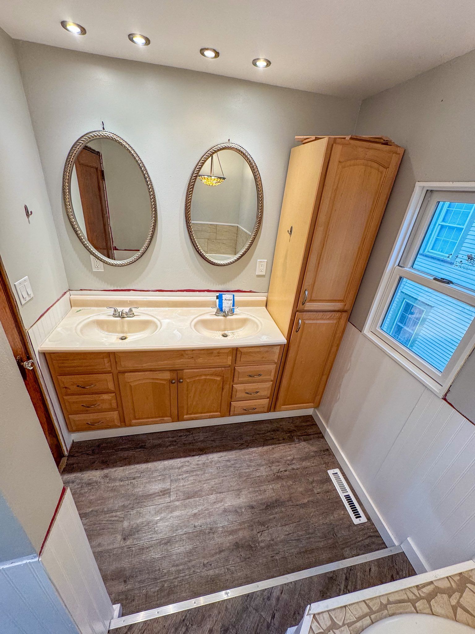 Bathroom with double vanity, oval mirrors, tall cabinet, and a window.