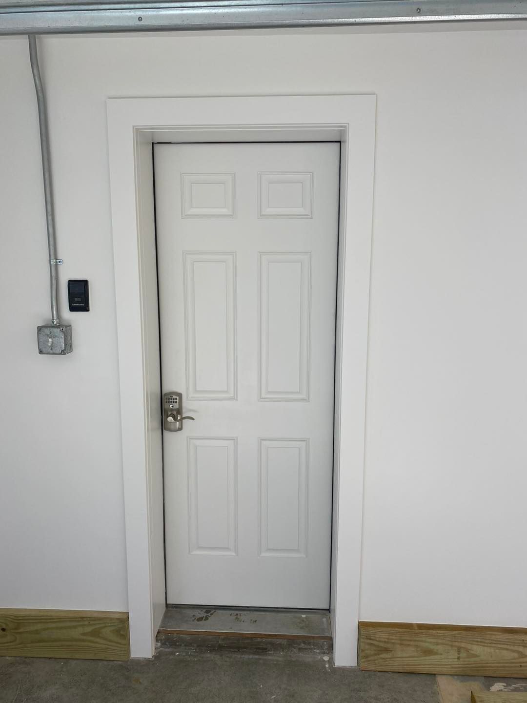 White six-panel door with molding, surrounded by white walls. Gray electrical conduit runs up the wall.