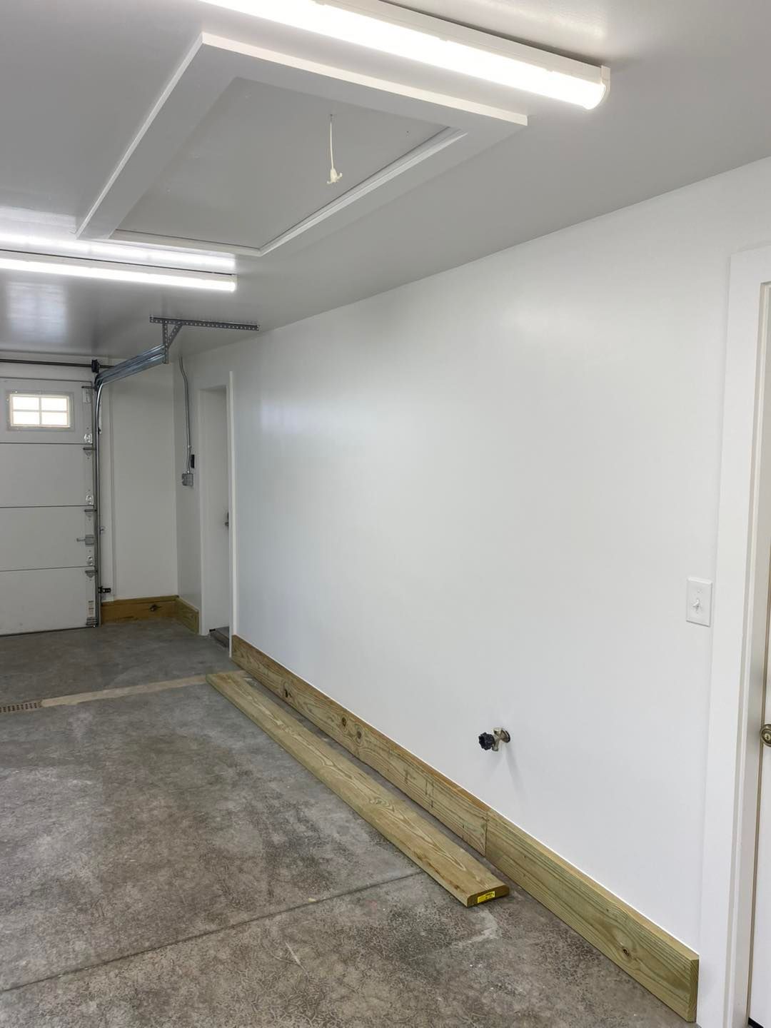 Garage interior with white walls, concrete floor, overhead lighting, and wooden planks along the wall.