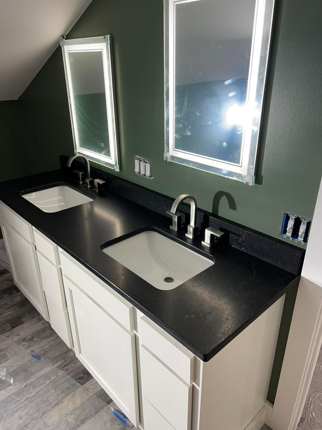 Bathroom with a white vanity, black countertop, two sinks, and mirrors. Dark green wall.