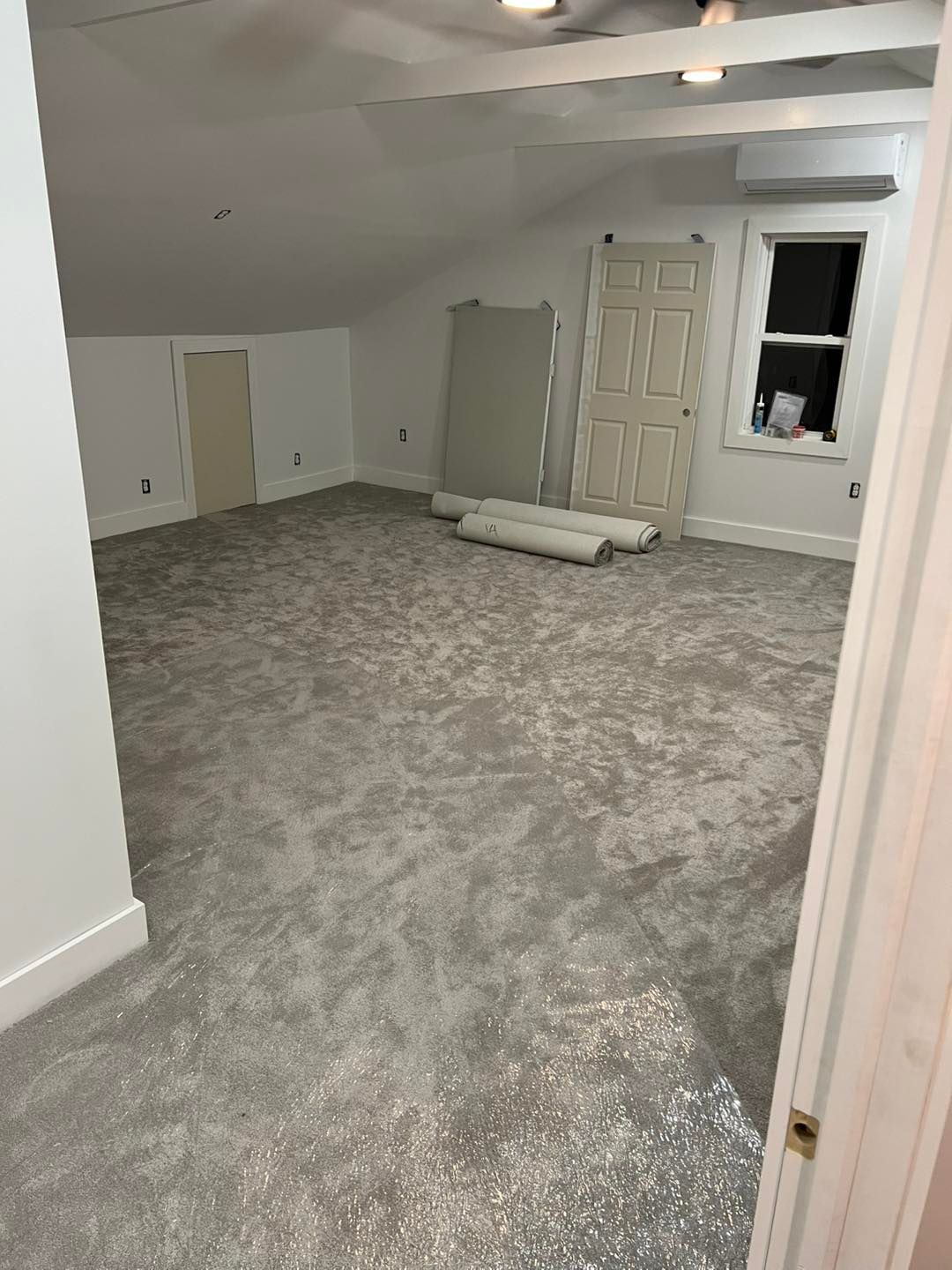 Room with new gray carpet, sloped ceiling, door, and window. Rolls of carpet and unfinished walls.