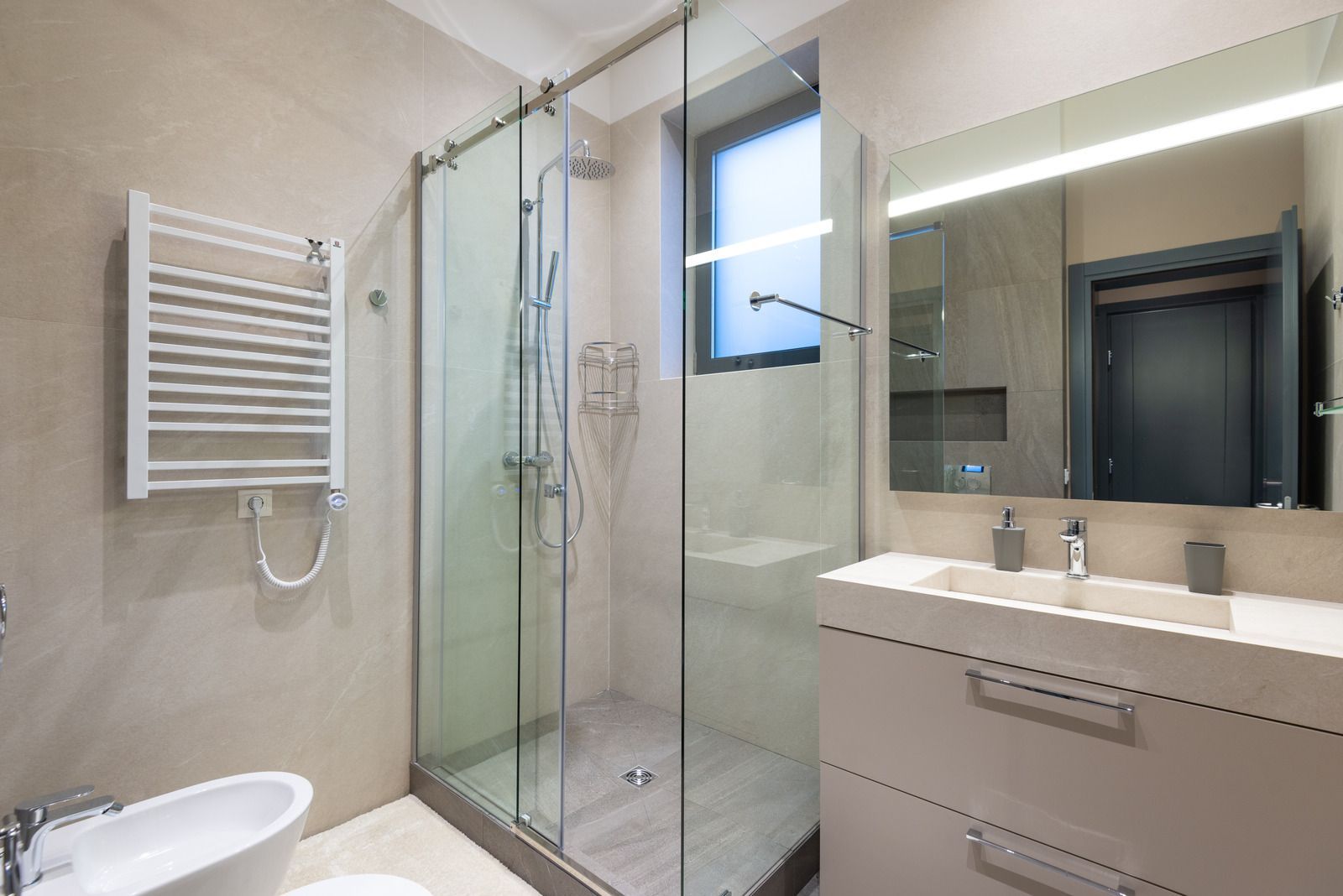 Modern bathroom with shower, vanity, heated towel rack, and bidet. Beige walls, gray shower floor, and a rectangular window.