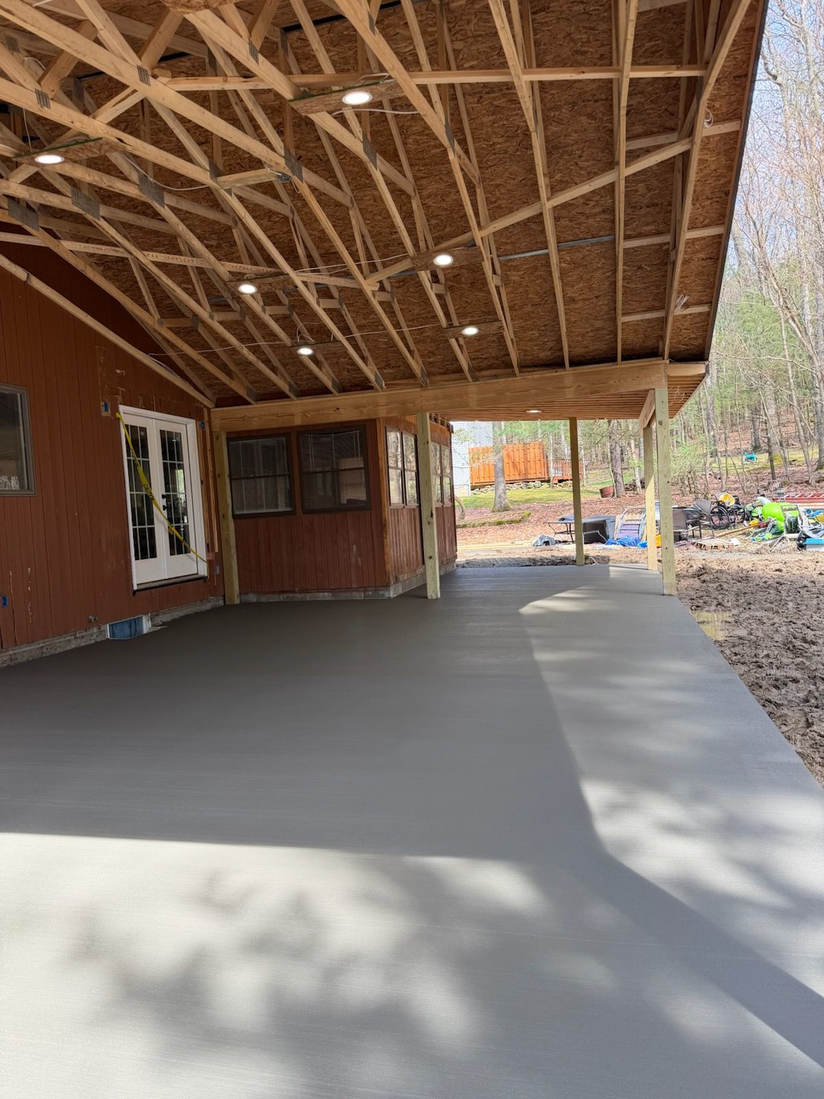 A new gray concrete patio covered by a wooden roof structure with recessed lighting, attached to a reddish-brown home.