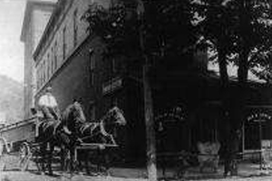 A black and white photo of a horse drawn carriage in front of a building.