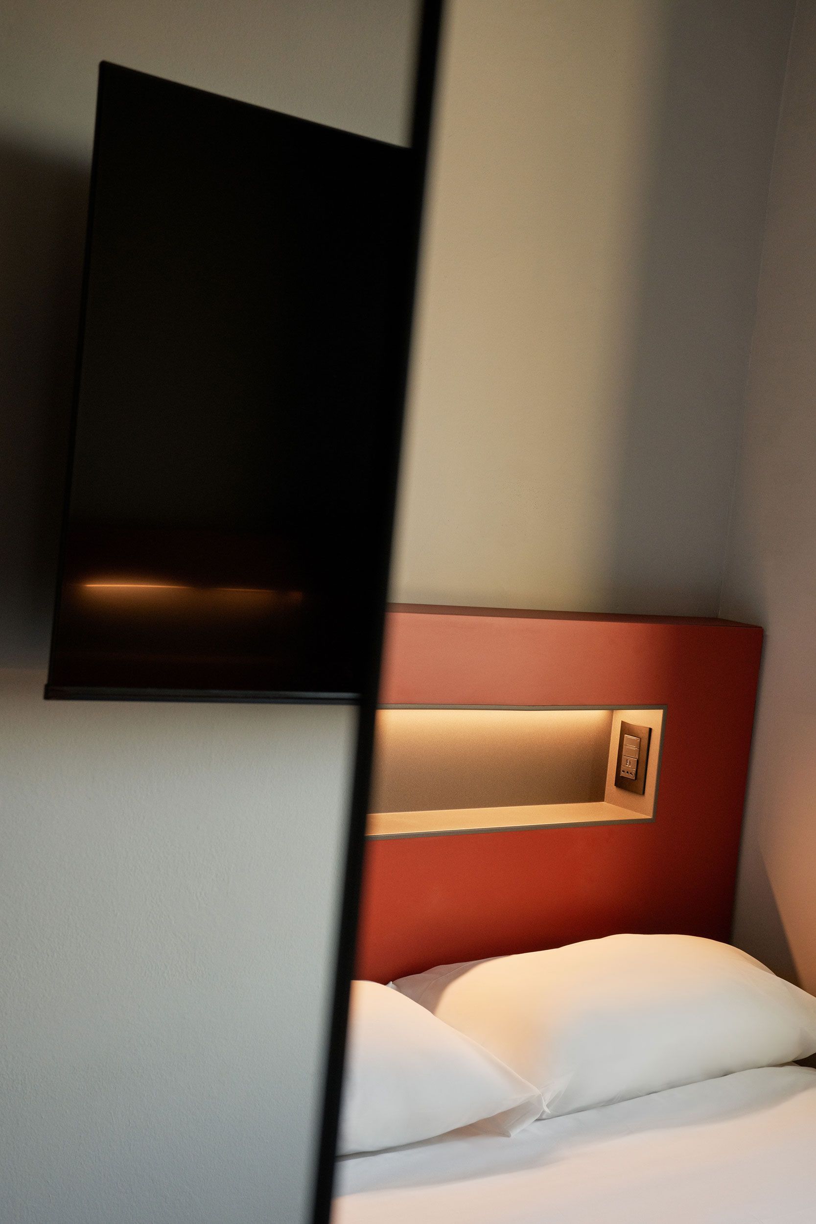 Una TV montata a parete sopra un letto con cuscini bianchi e una testiera rossa.