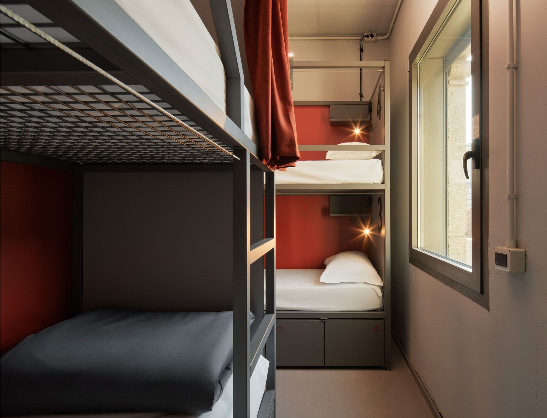 Letti a castello in una stanza d'ostello. Tre letti sovrapposti con dettagli rossi e una finestra.