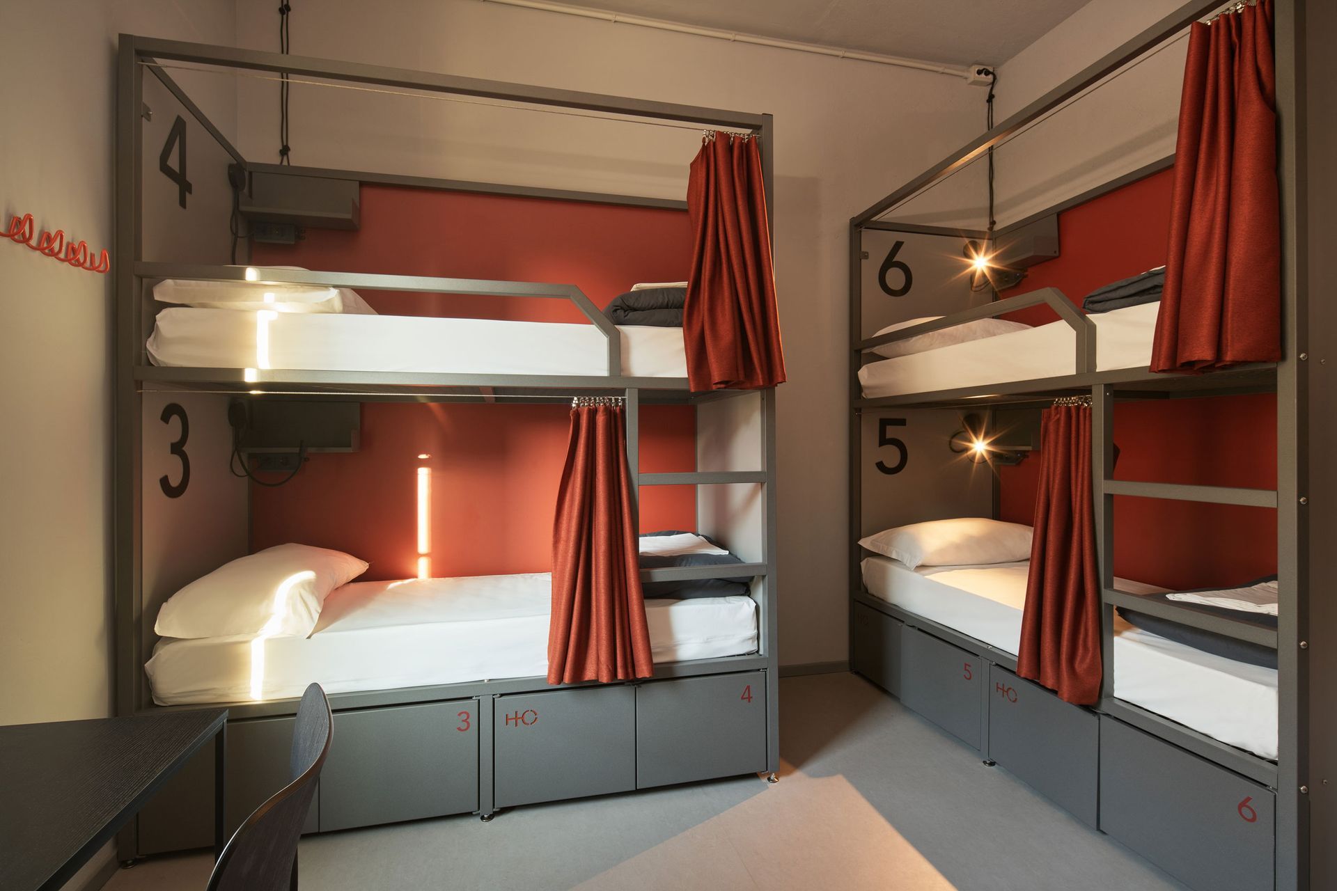 Letti a castello in una stanza d'ostello con tende arancioni e armadietti grigi. Sono visibili i numeri 3, 4, 5 e 6.