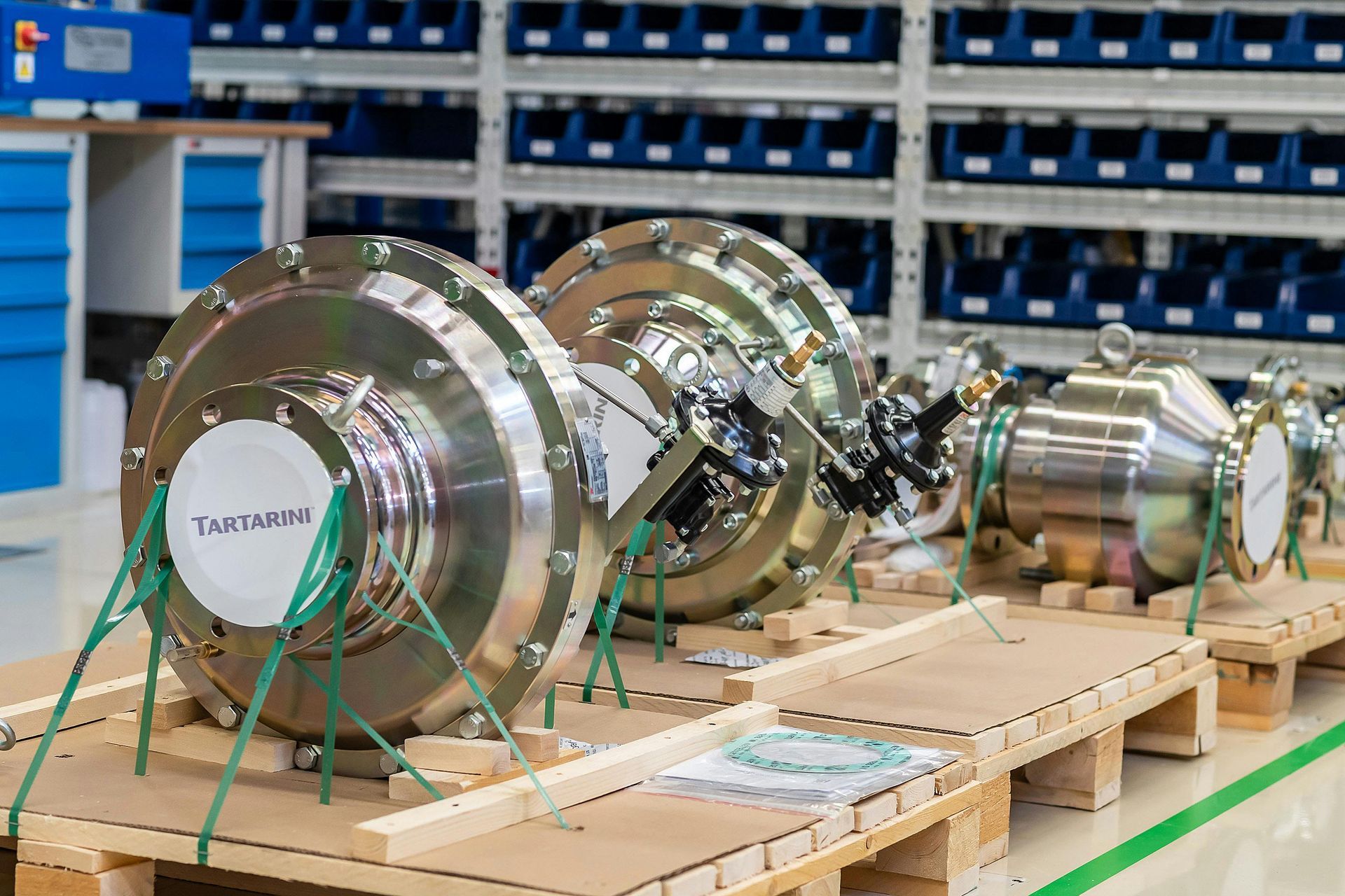 Three shiny, cylindrical industrial components on wooden pallets in a warehouse with shelves of blue bins.