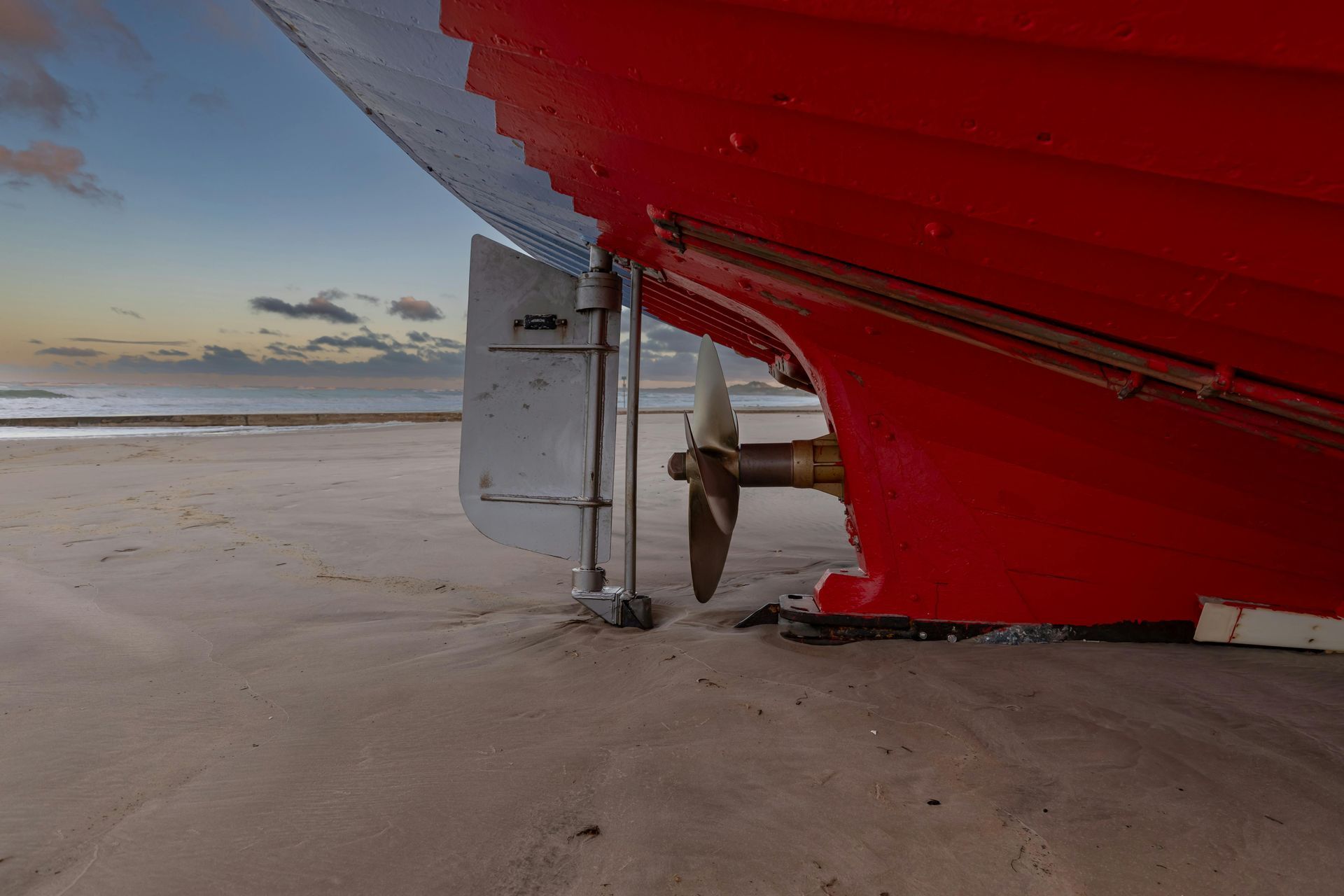 Red and blue boat on a beach, with propeller visible, setting sun in background.