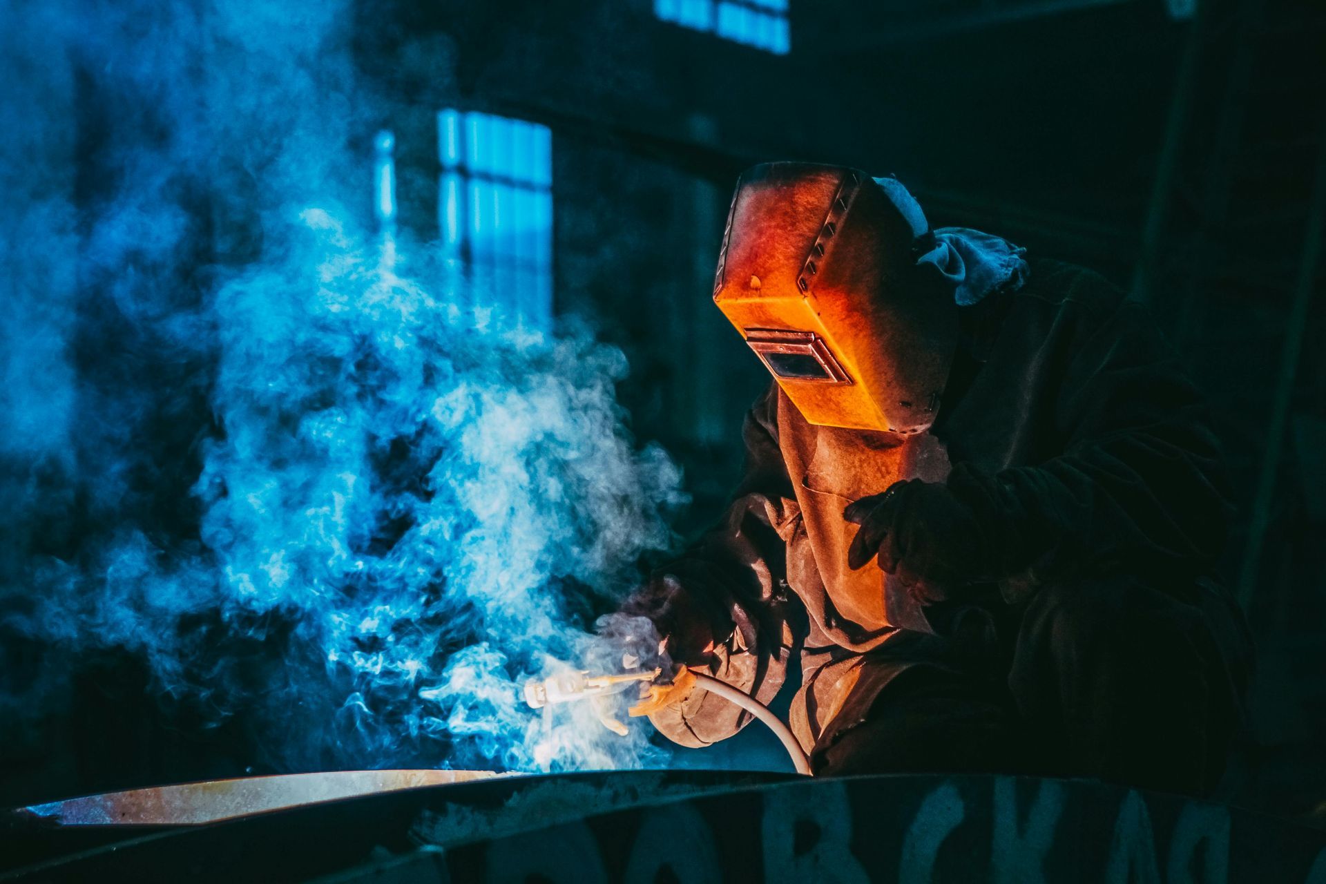 Welder in orange helmet at work, sparks flying, surrounded by blue smoke in a dark workshop.