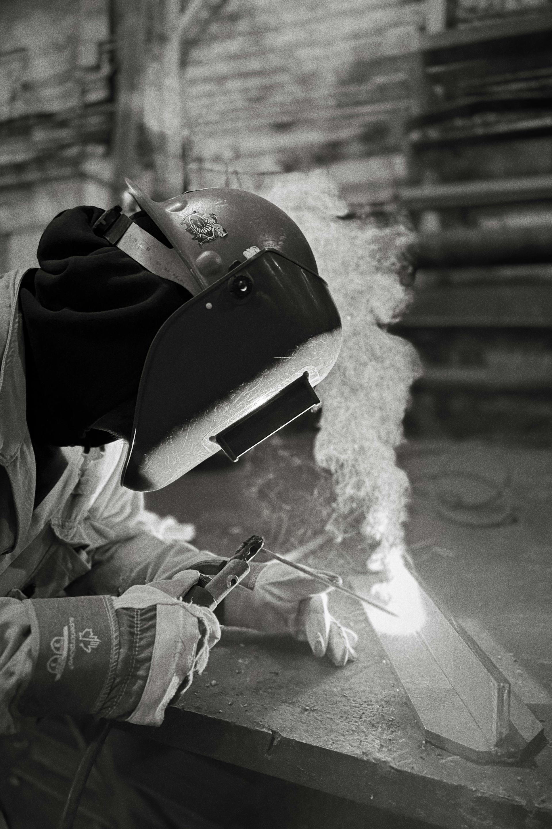 Welder wearing protective gear, welding metal, with sparks and smoke in a workshop setting.