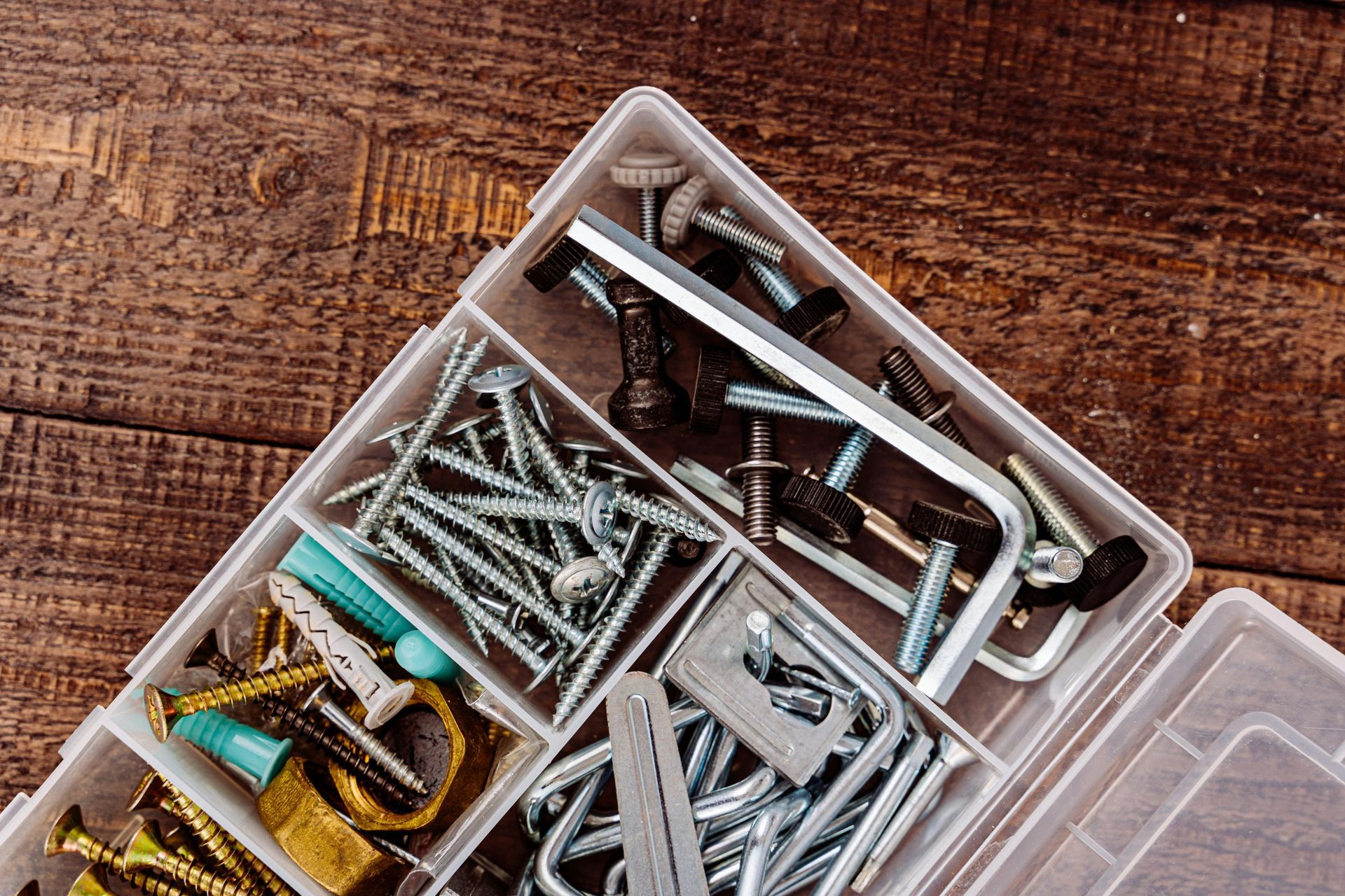 Open plastic organizer filled with various screws, bolts, and tools on a wooden surface.