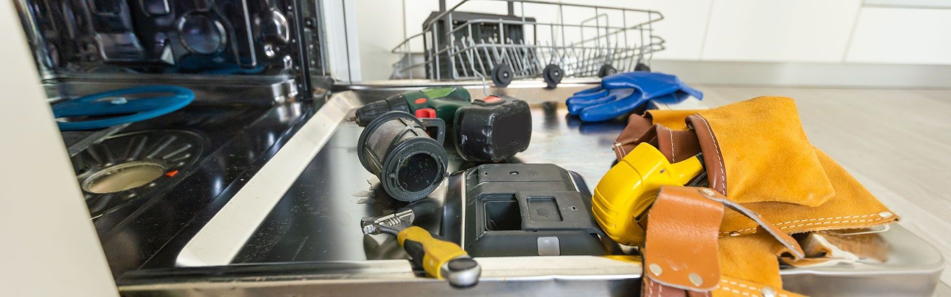 A dishwasher is being repaired in a kitchen with tools and a tool bag.