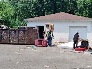 A group of people are standing in front of a dumpster in a parking lot.