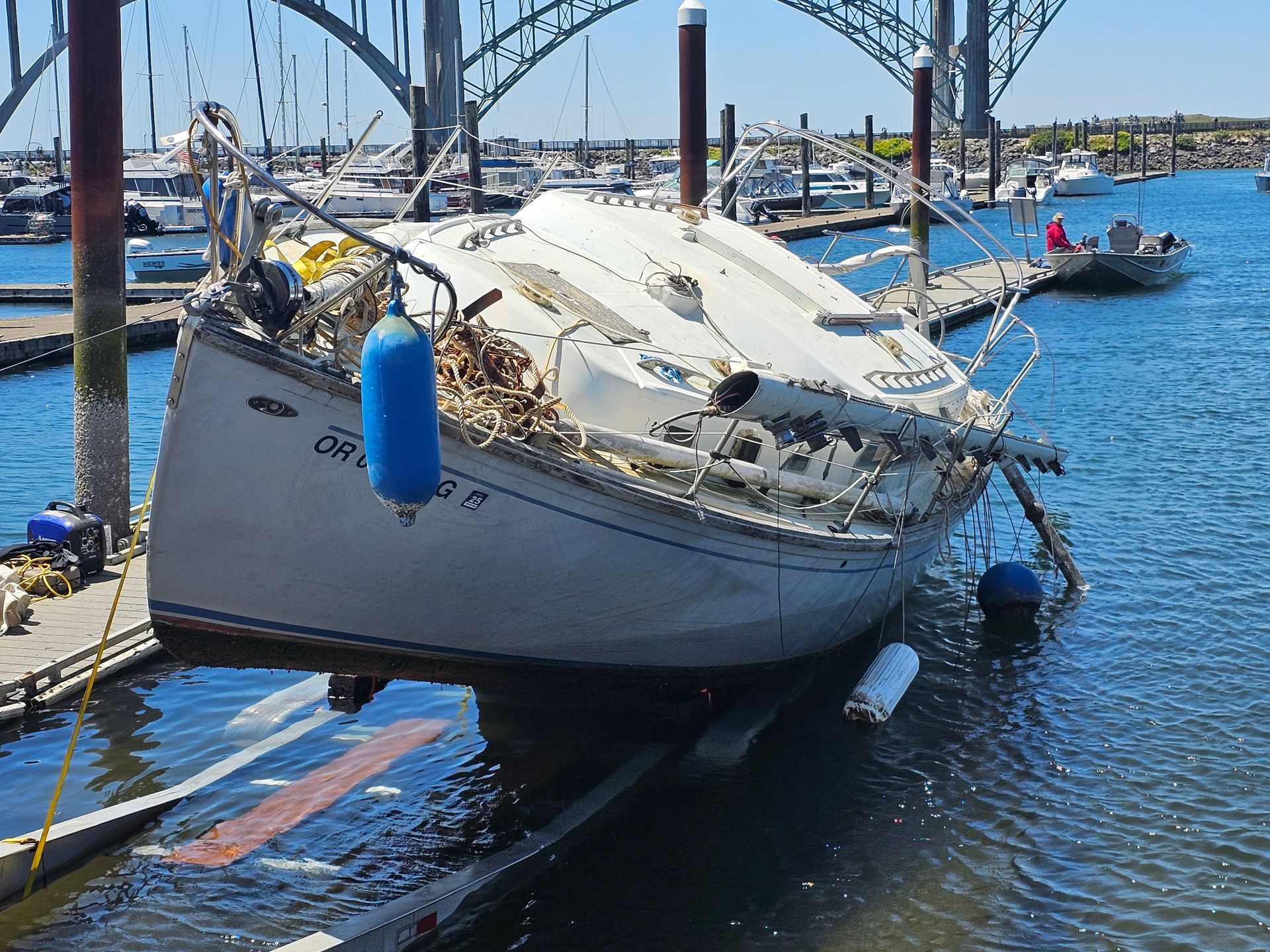 A damaged sailboat sits on a ramp at a marina, under a bridge. Blue water and sky.