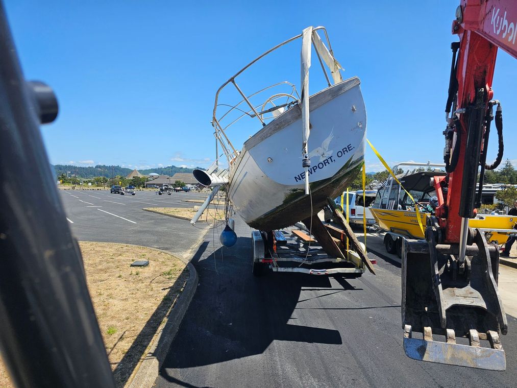 A boat on a trailer being moved by an excavator on a sunny day in a parking lot.