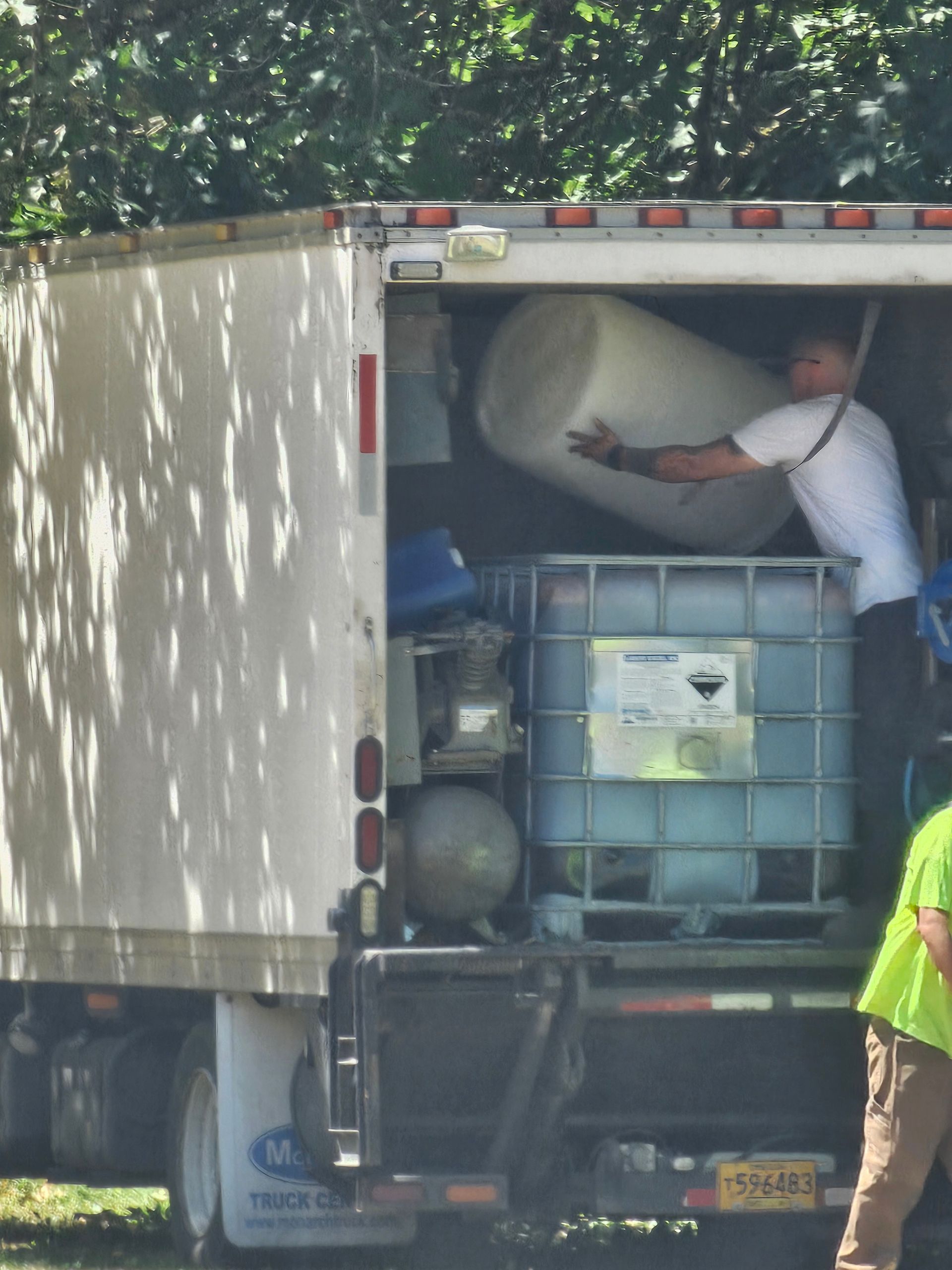 Man loading a large cylindrical tank into a truck, next to a large container. Outdoor setting.