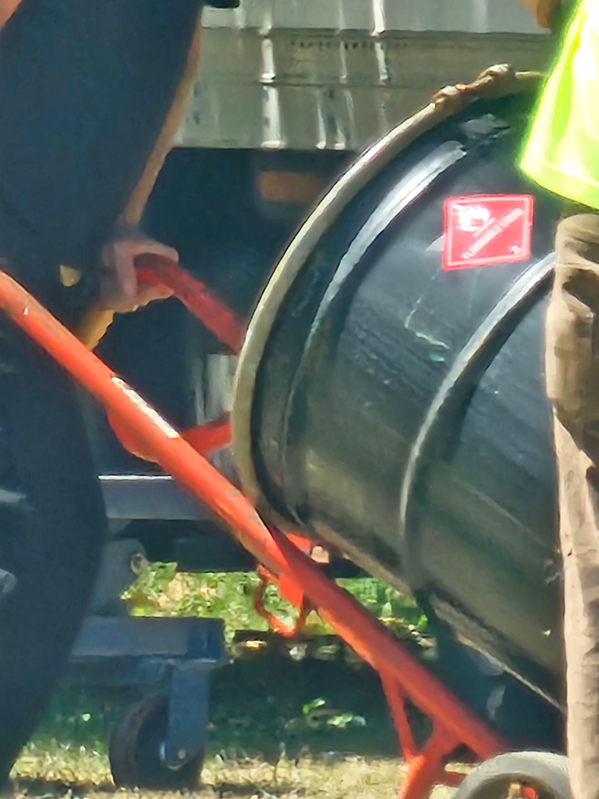 Black barrel with hazard symbol on an orange hand truck, being maneuvered by people outdoors.
