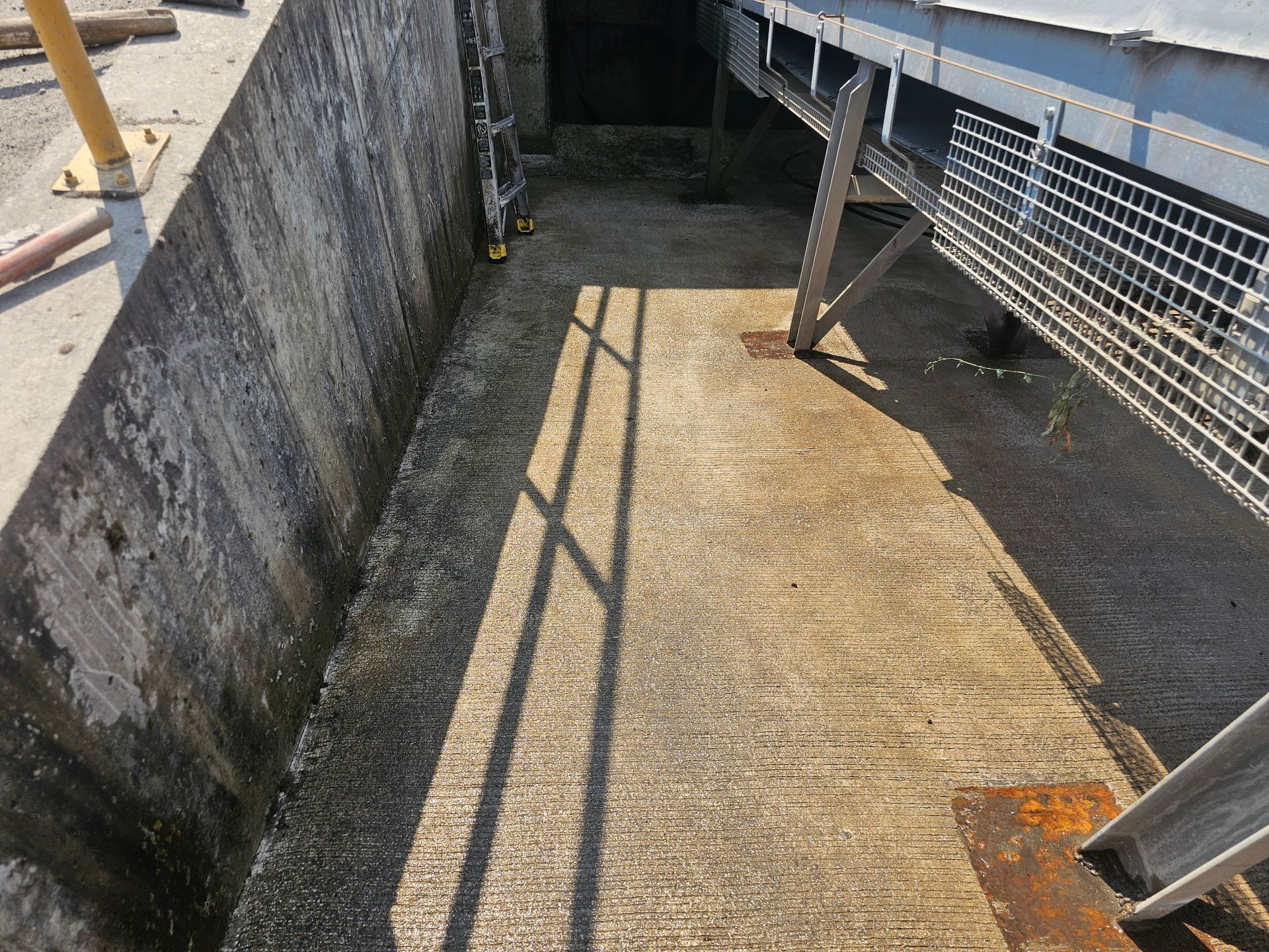 Concrete walkway next to a cement wall and a conveyor belt structure, with sunlight casting shadows.