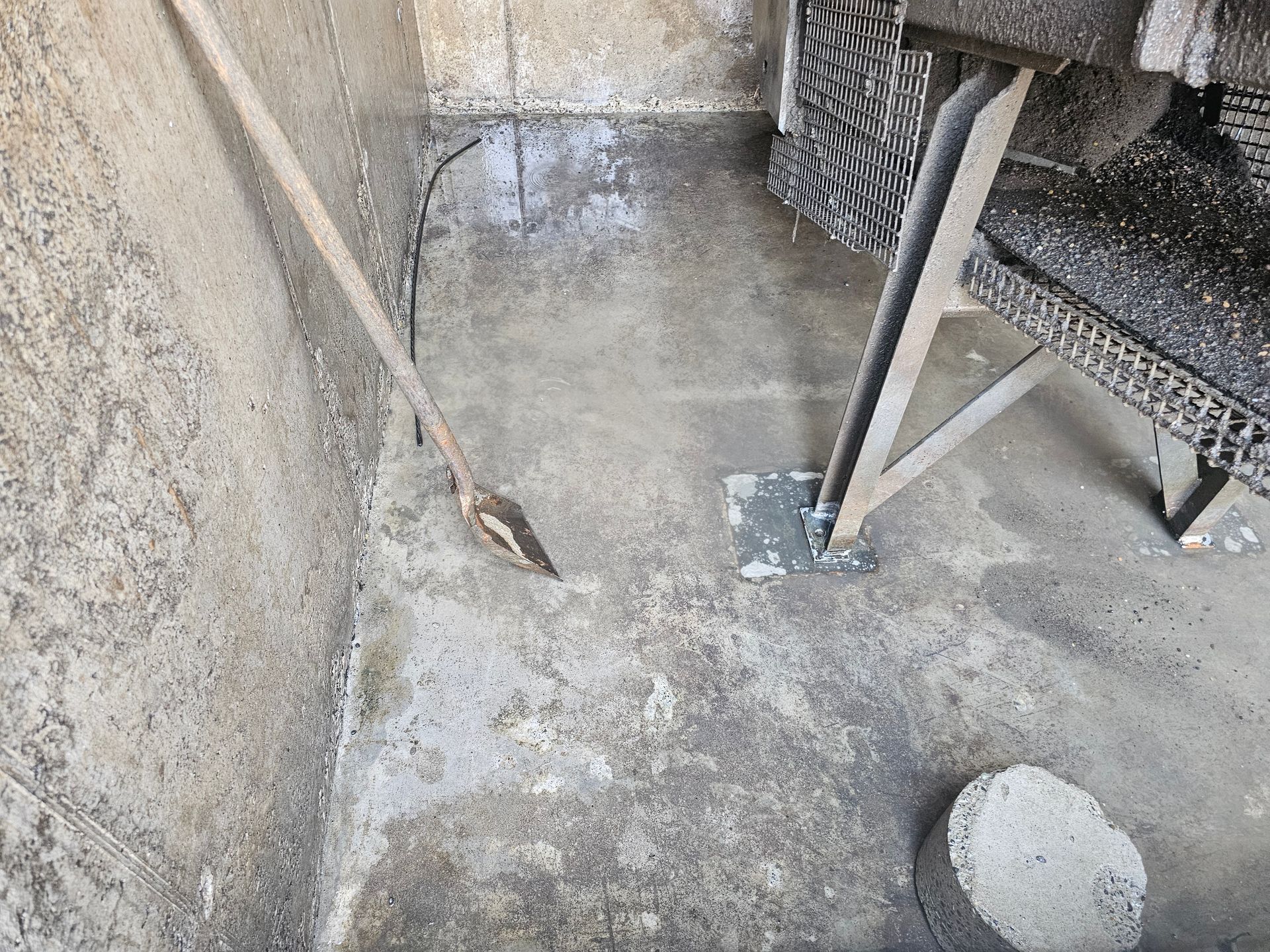 Concrete basement corner with a shovel, a metal shelf, and wet cement floor.