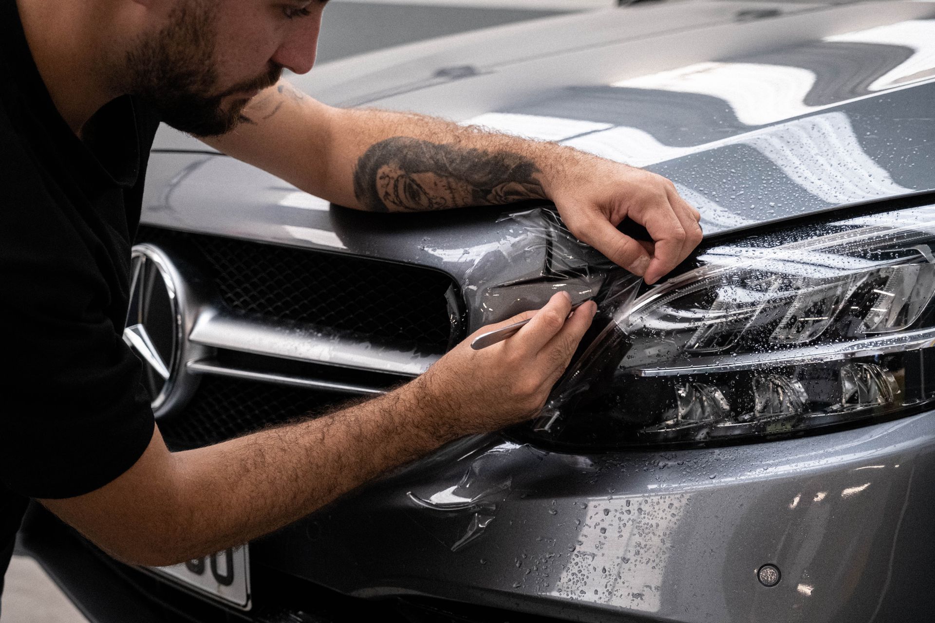 Person applying tint to a car headlight. Gray car, black tint, and person with tattoos.