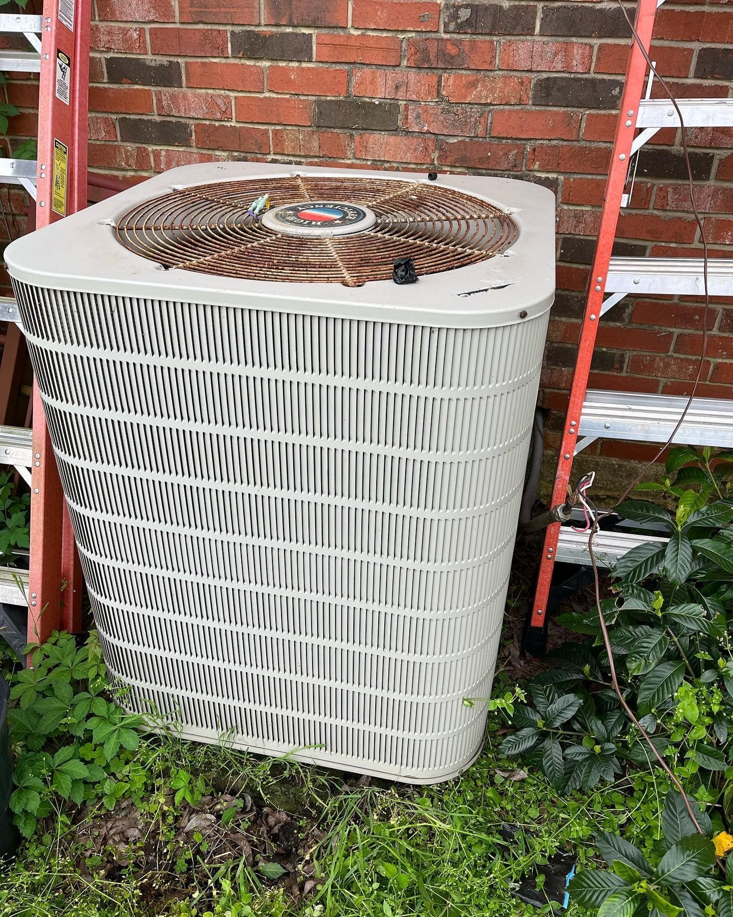 A large air conditioner is sitting in front of a brick wall next to a ladder.