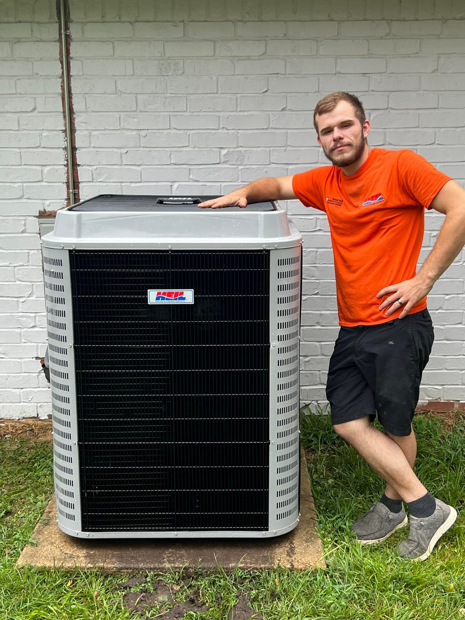 A man in an orange shirt is standing next to a large air conditioner.